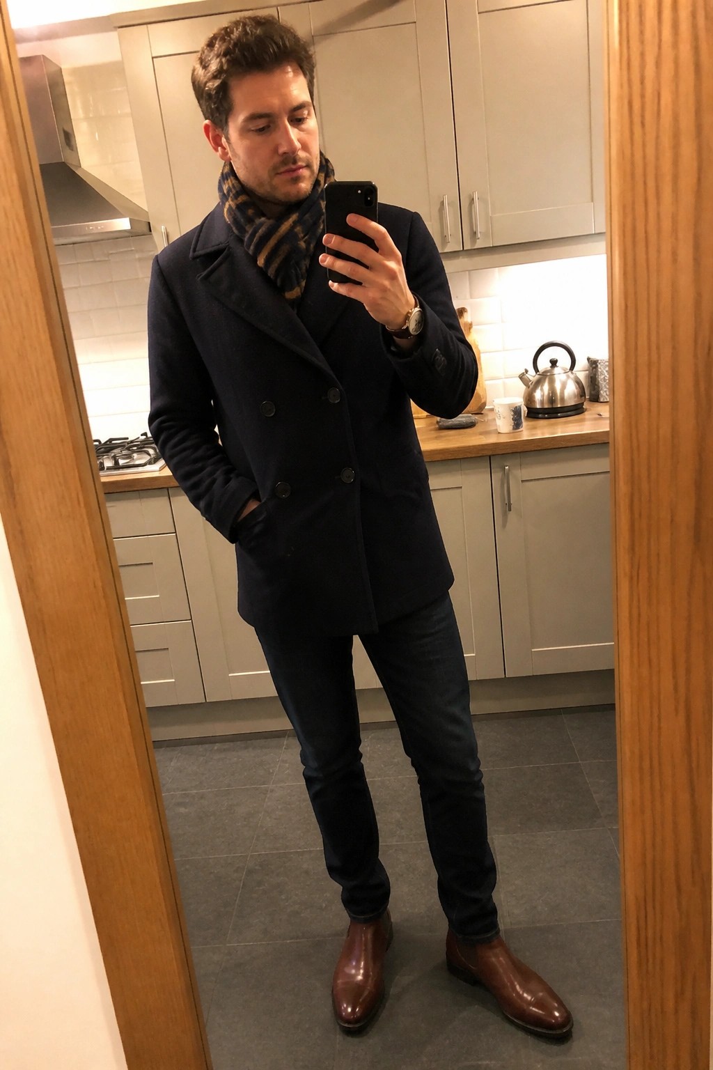 Man in navy peacoat and plaid scarf taking kitchen mirror selfie