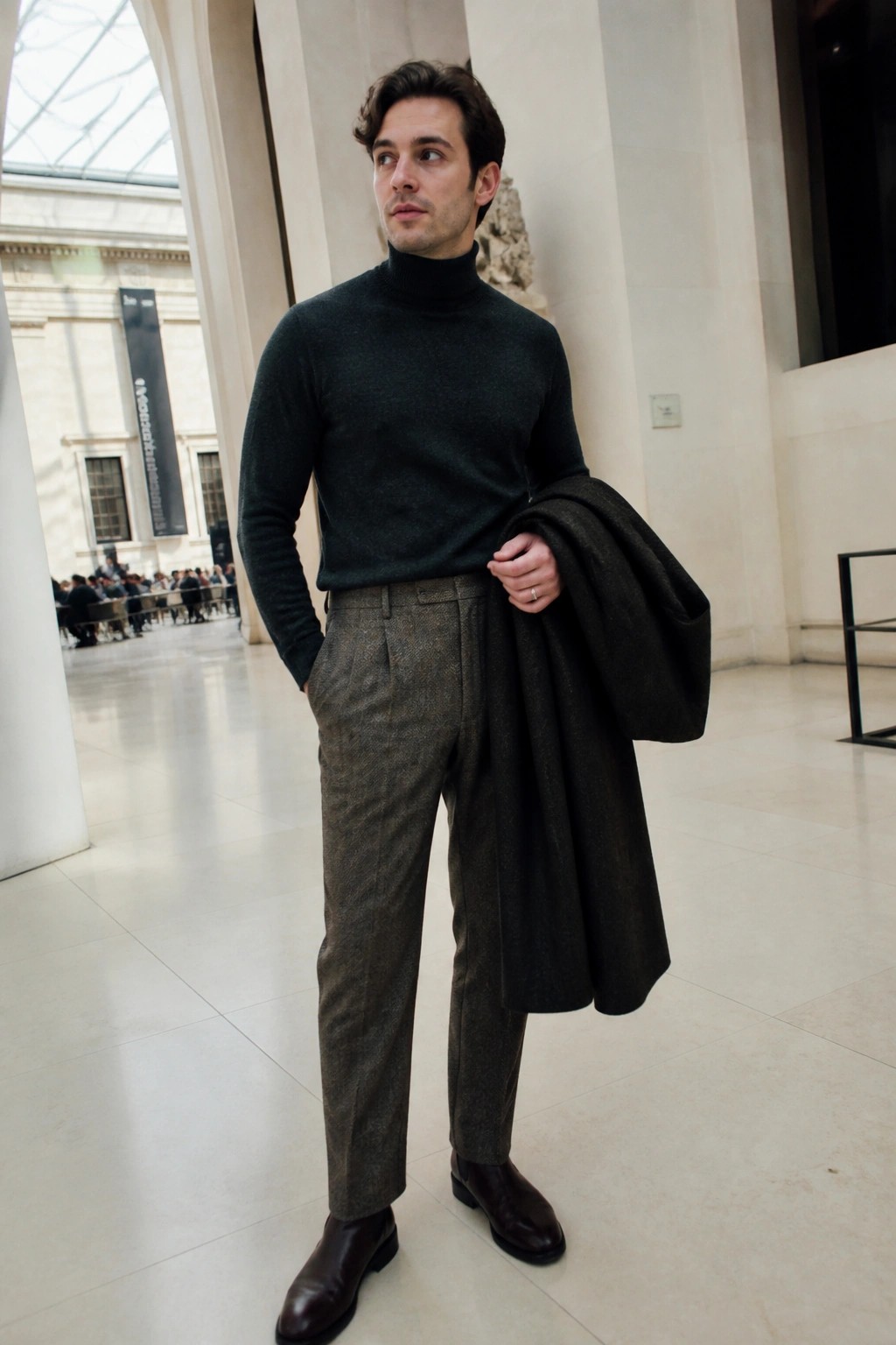 Man in dark green turtleneck and tweed trousers holding coat in marble hall.