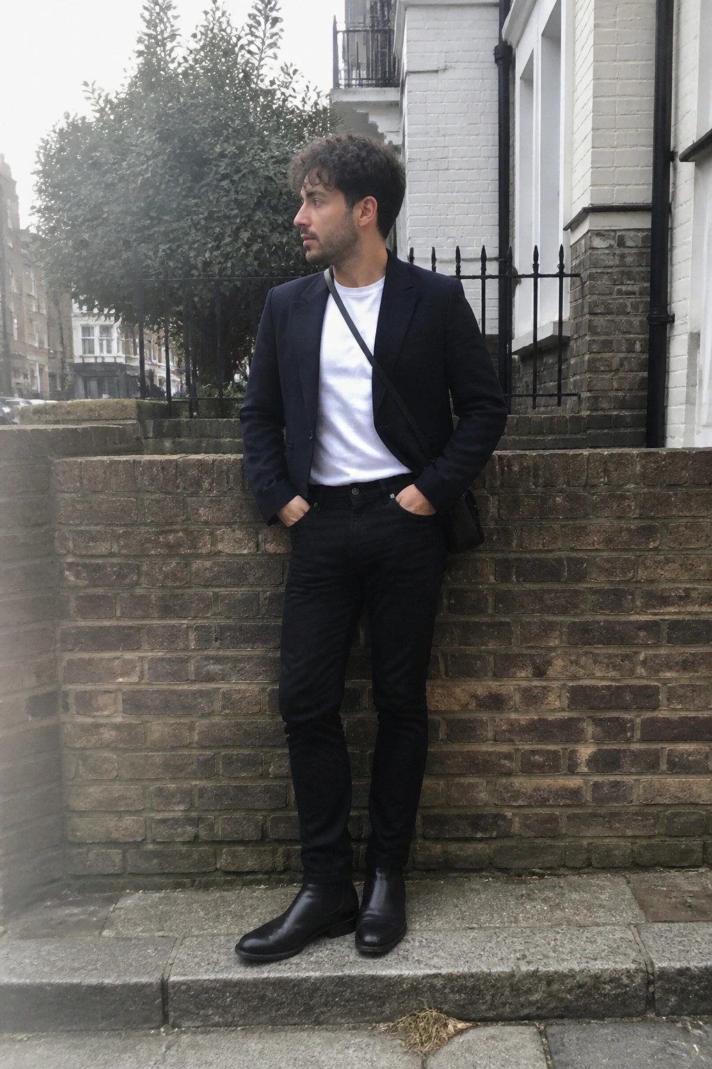 Man with curly hair in navy blazer over white t-shirt and black jeans by brick wall