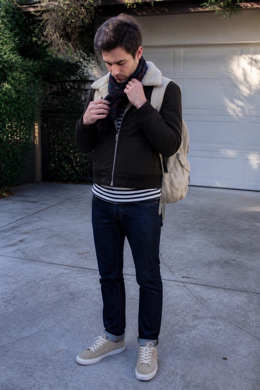 Man in brown shearling jacket adjusts plaid scarf with backpack on driveway