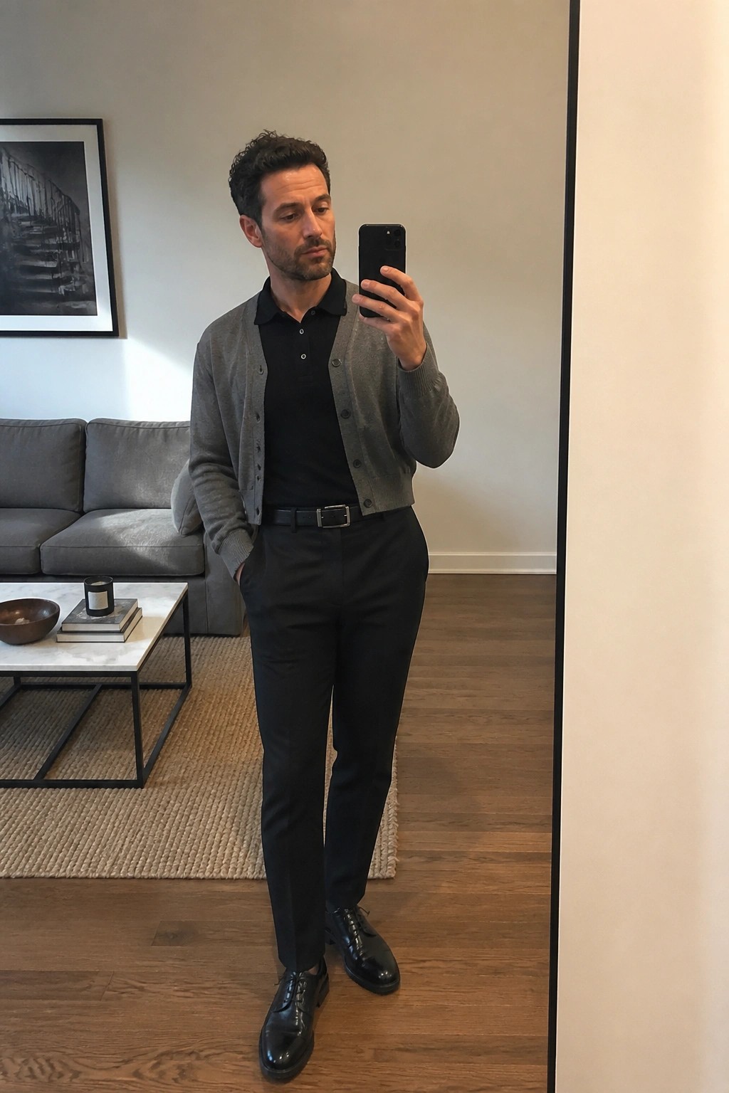 Man in gray cardigan over black polo taking mirror selfie in living room