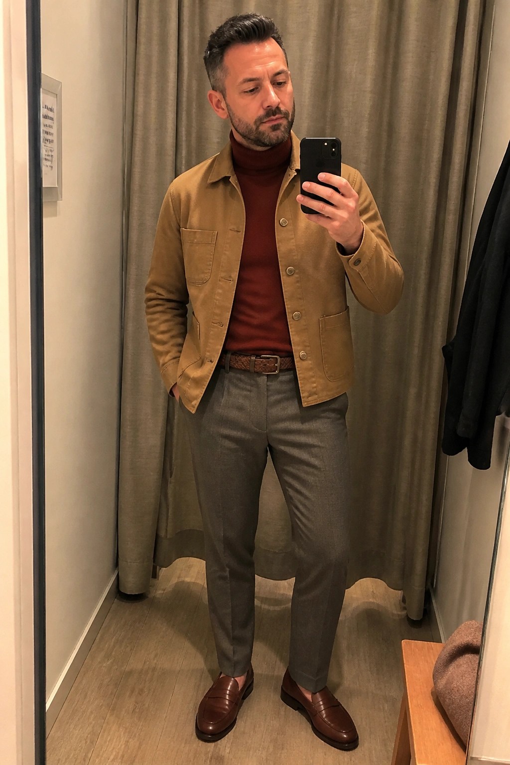 Man in tan jacket over maroon turtleneck, gray trousers, and brown loafers taking mirror selfie