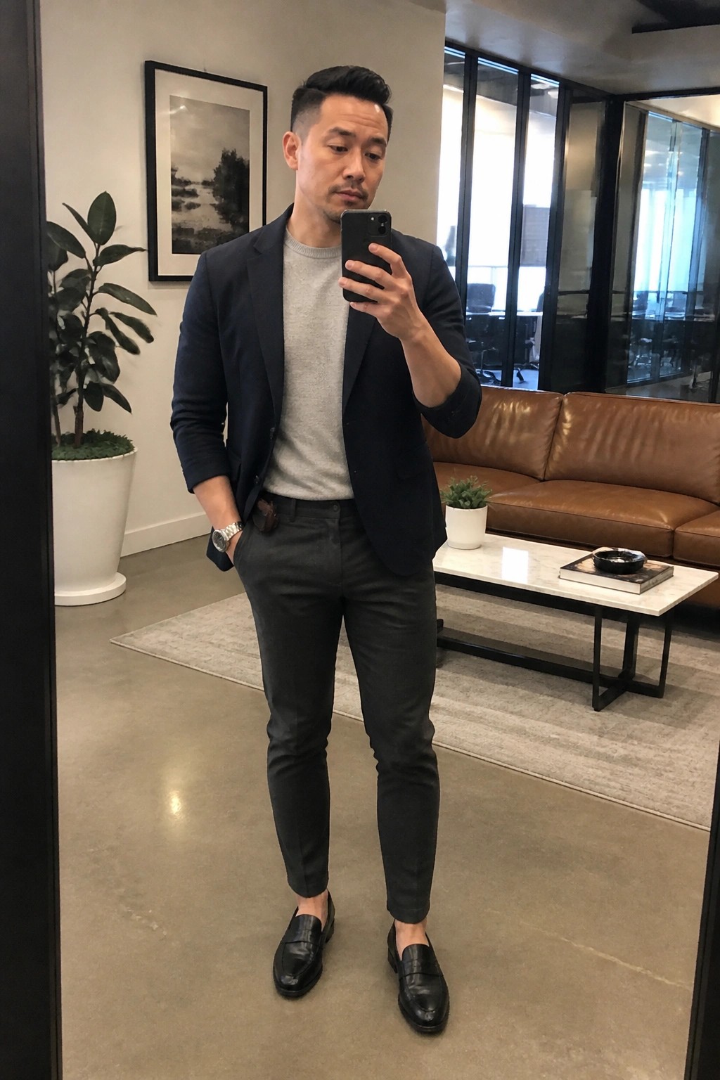 Man in navy blazer over gray sweater takes mirror selfie in modern lounge.