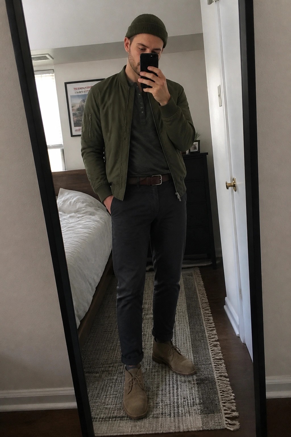 Man in green bomber jacket and beanie taking mirror selfie in bedroom