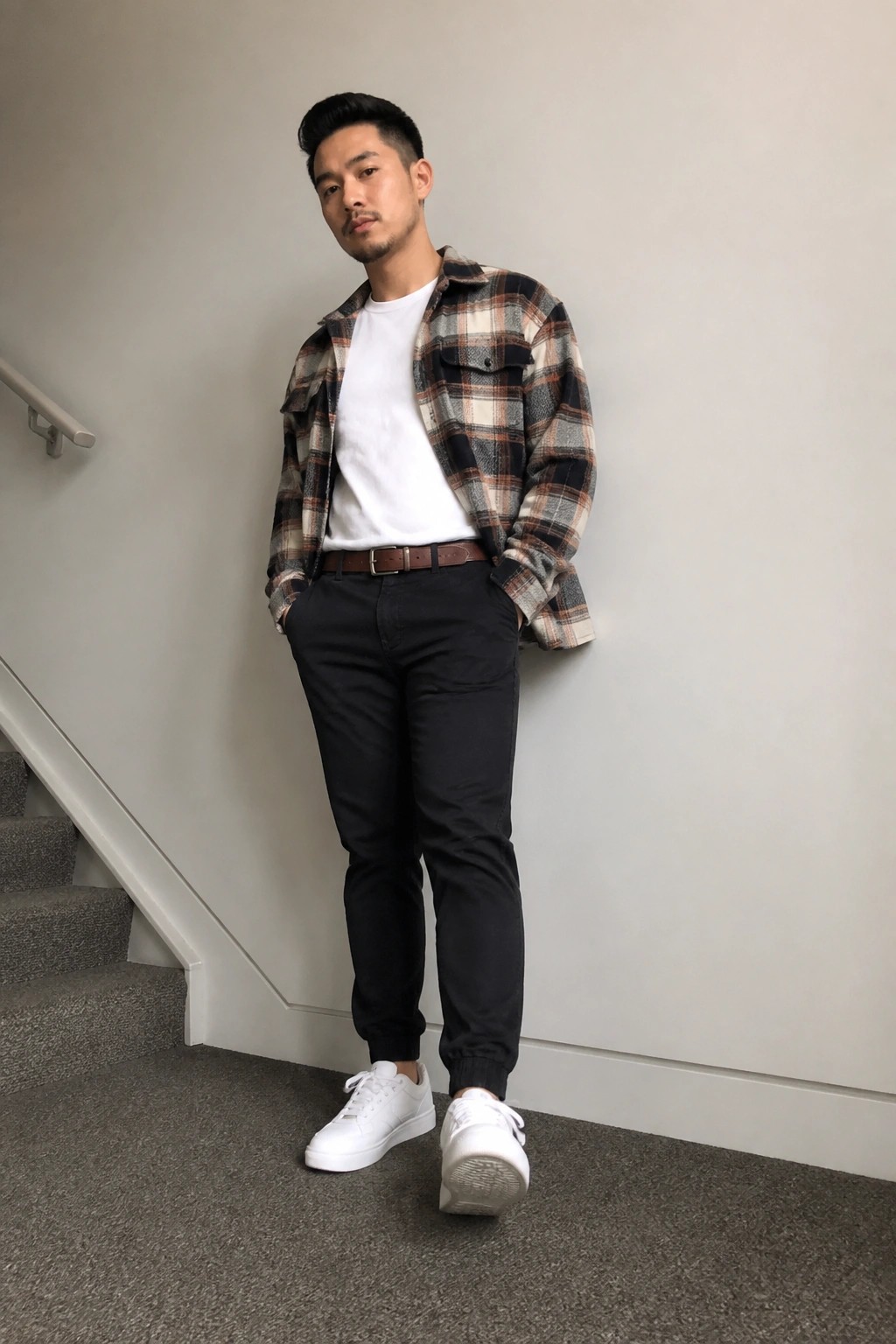 Man in plaid flannel shirt over white tee, black pants, and white sneakers by stairs