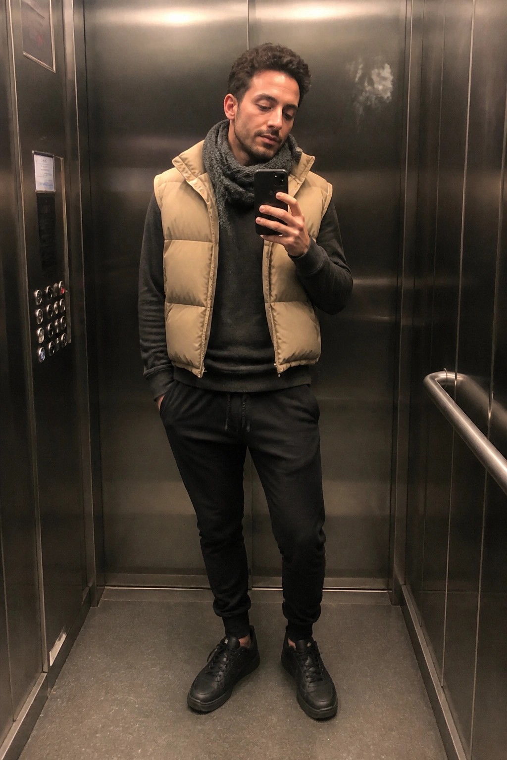 Man in beige puffer vest and gray scarf holds phone in elevator.