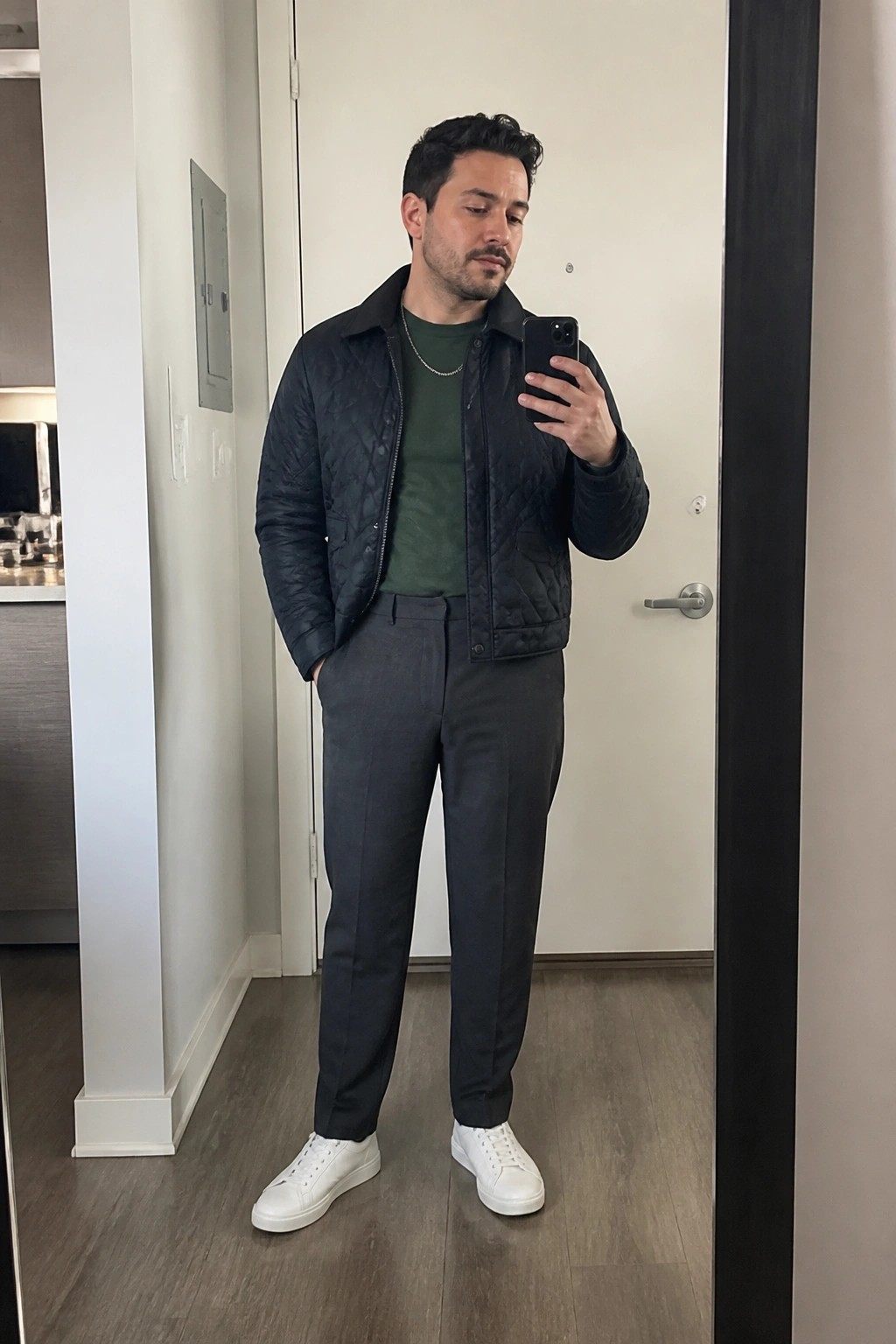 Man in dark quilted jacket over green shirt taking mirror selfie in gray pants