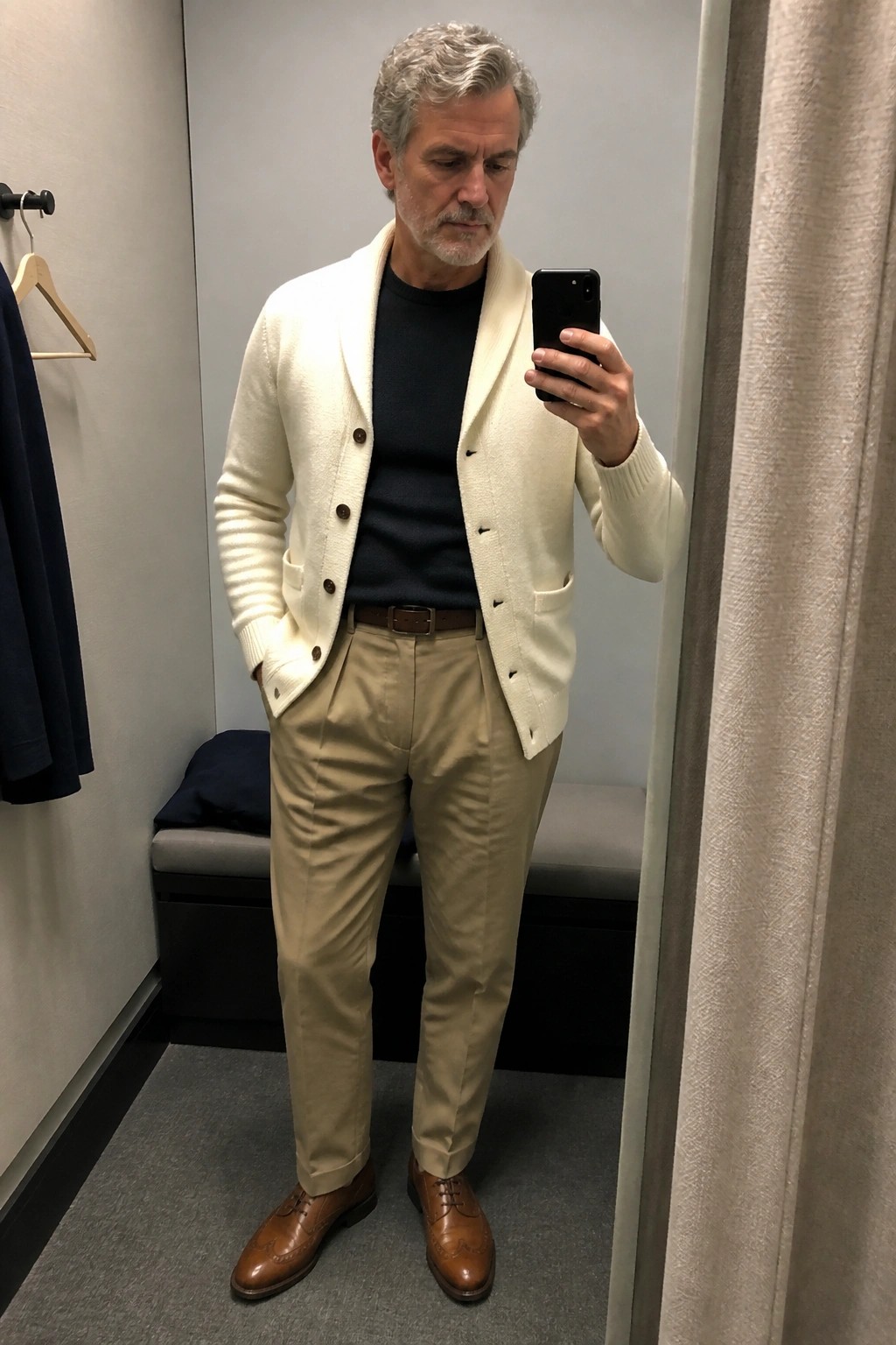 Man with gray hair in cream cardigan taking mirror selfie in fitting room