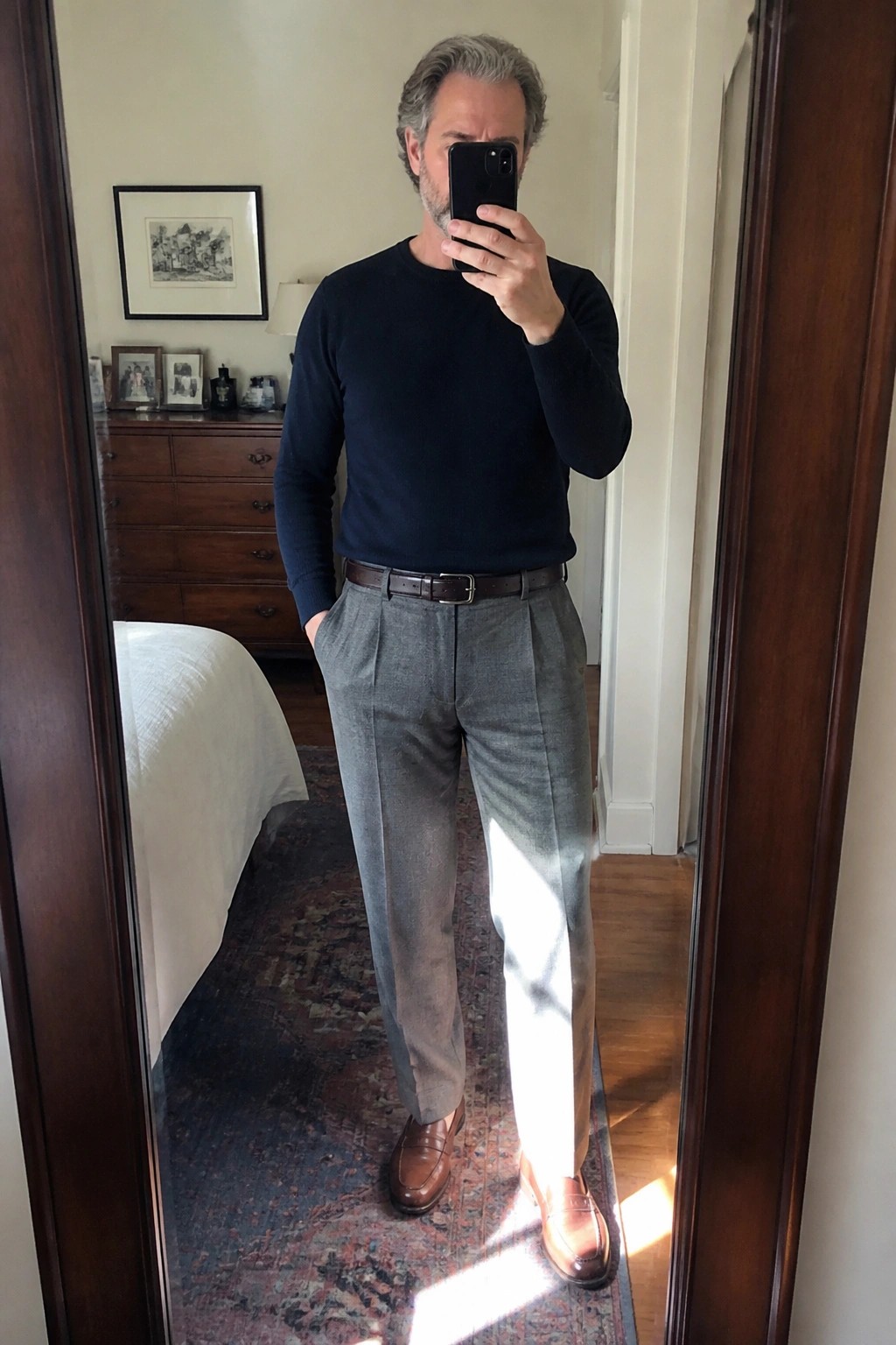 Man in navy sweater and gray pleated trousers taking mirror selfie with phone