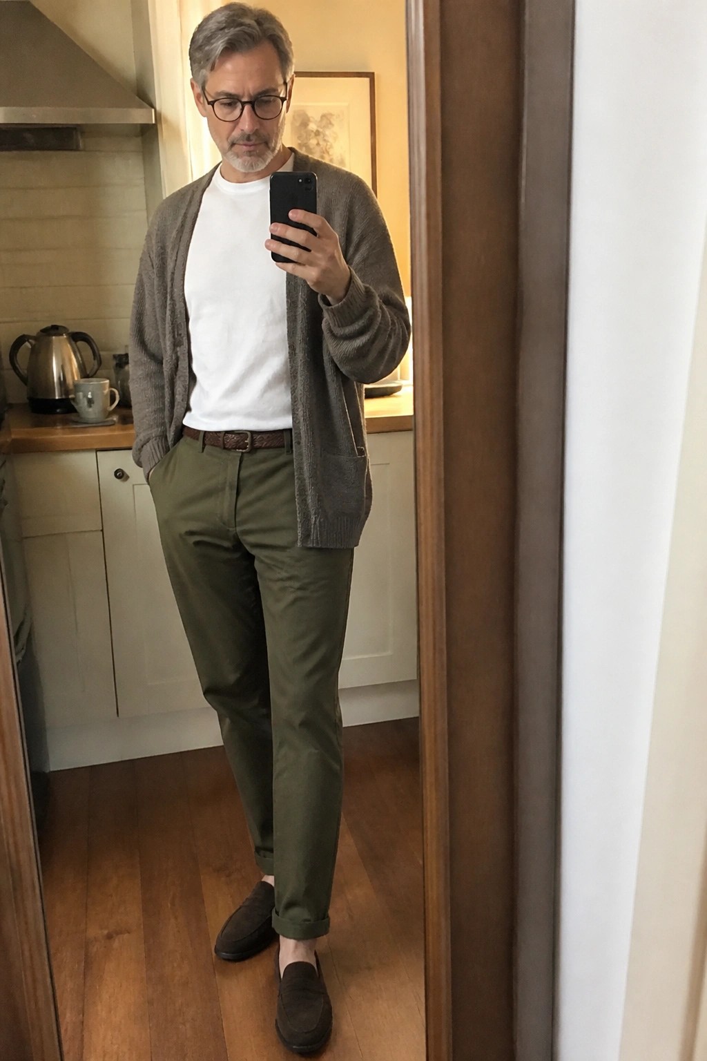 Man in gray cardigan and olive pants taking mirror selfie with phone