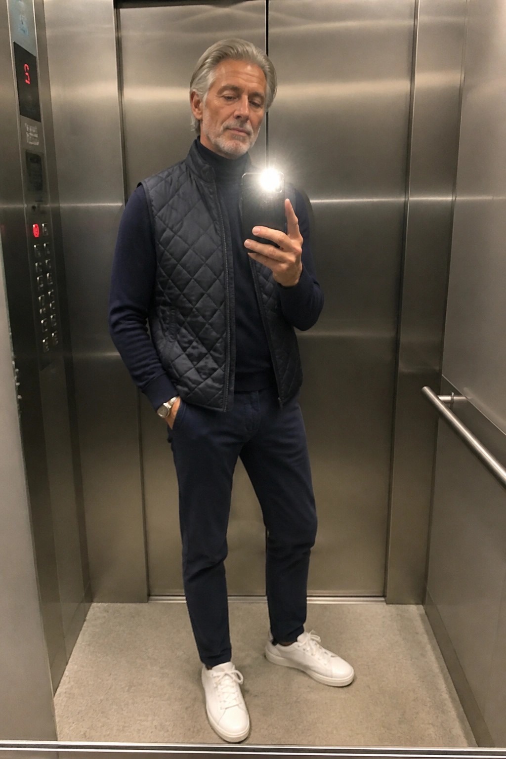 Gray-haired man in navy quilted vest taking flash selfie in elevator