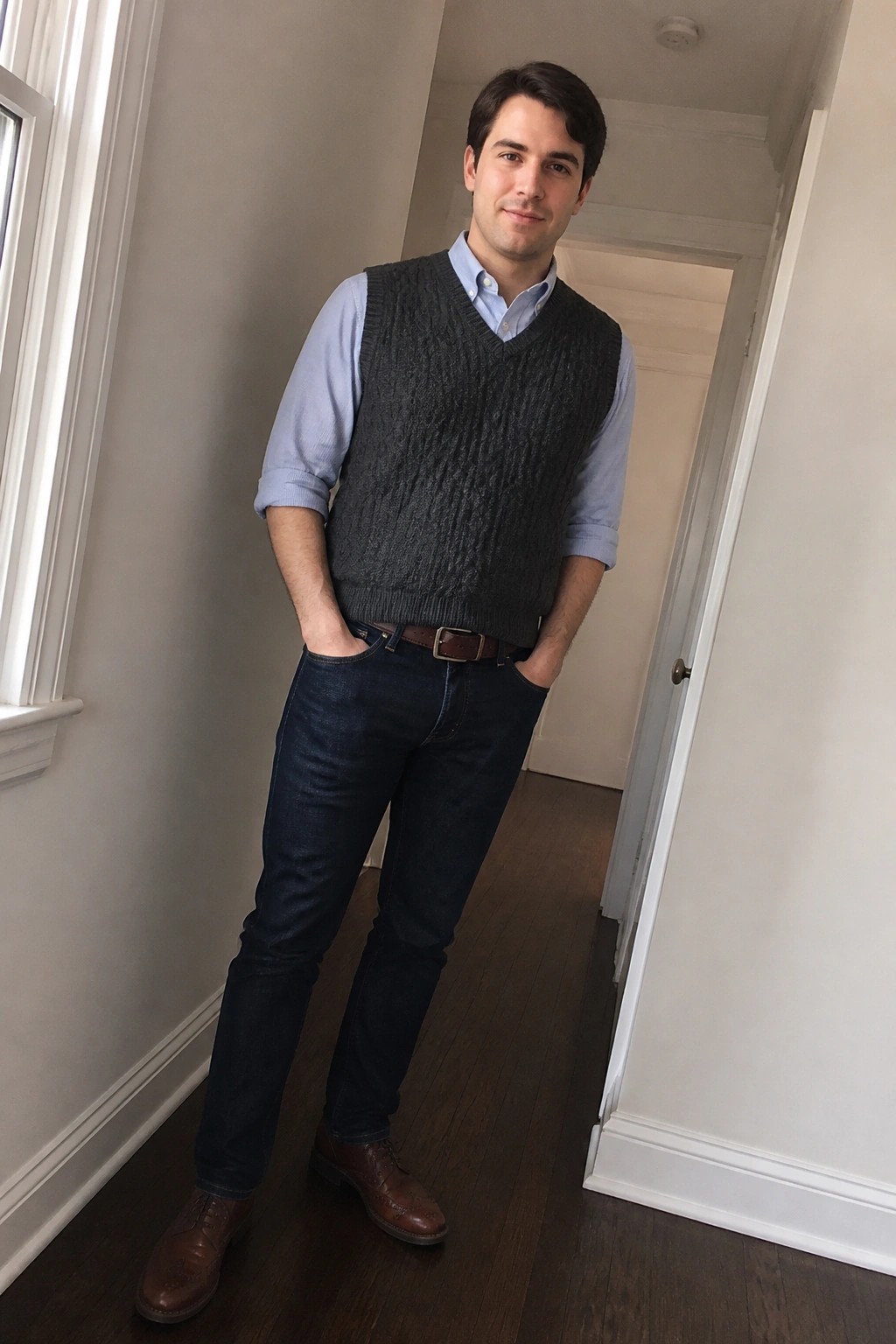 Man wearing a light blue button down shirt under a gray cable knit sweater vest, dark blue jeans, brown leather belt, and brown brogue shoes with hands in pockets.