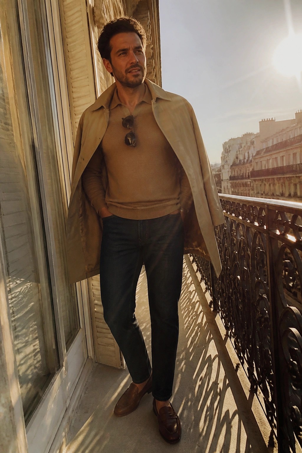 Man in beige trench coat over brown sweater on sunny Paris balcony.