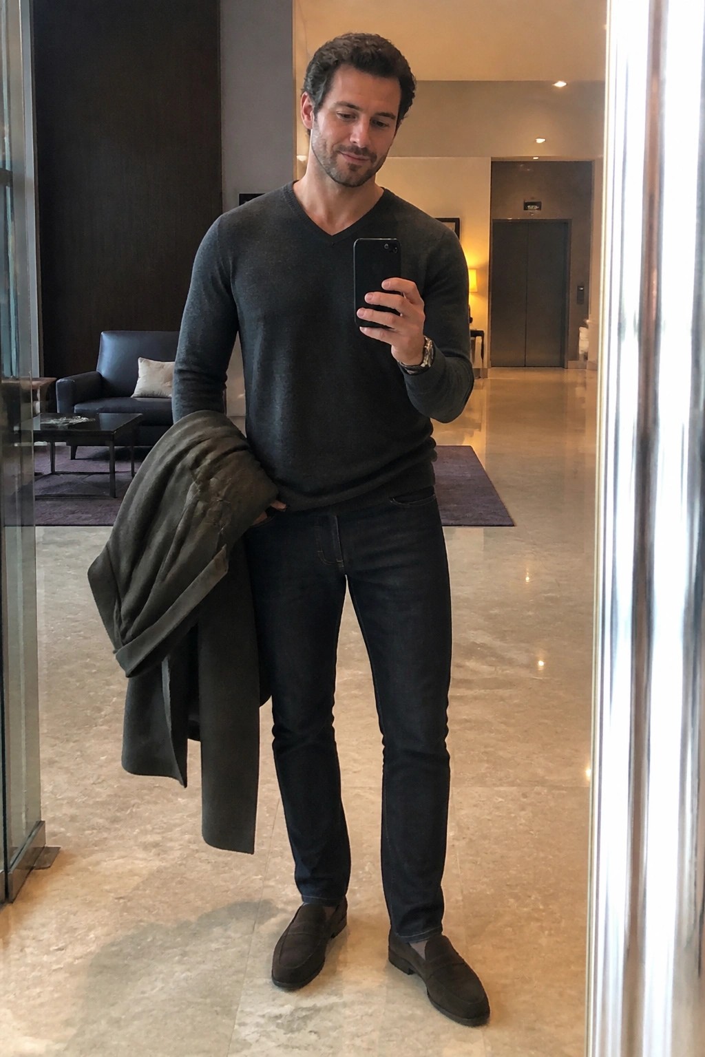 Man in gray sweater taking mirror selfie while holding dark jacket in lobby