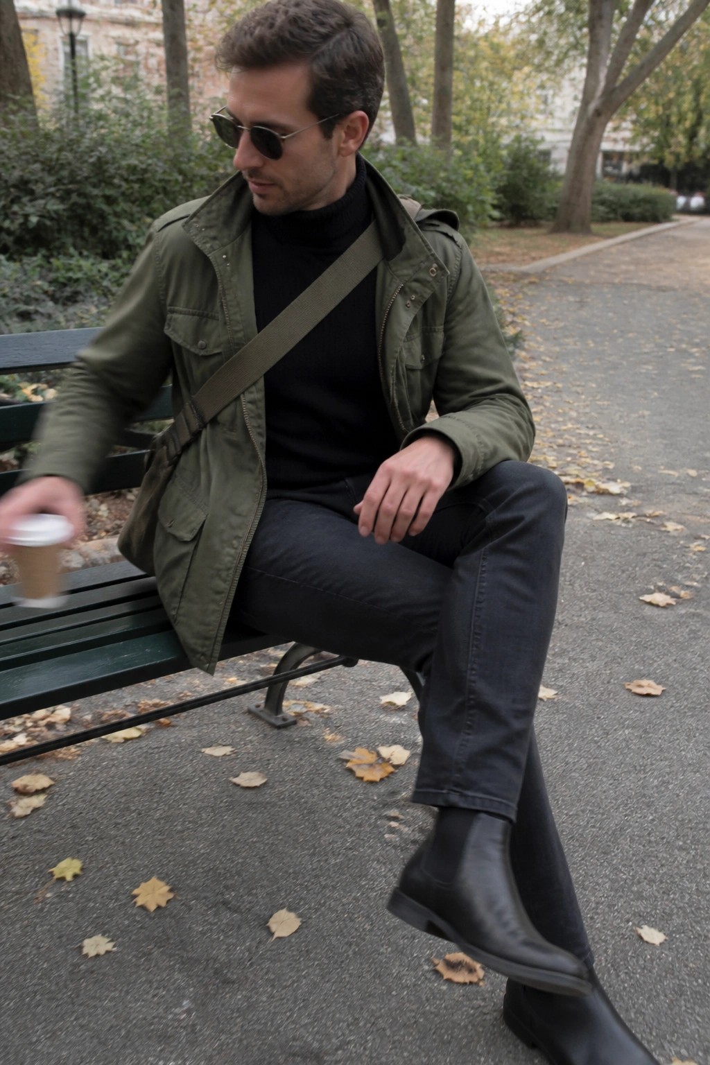 Man in green jacket and black boots on autumn park bench holding coffee