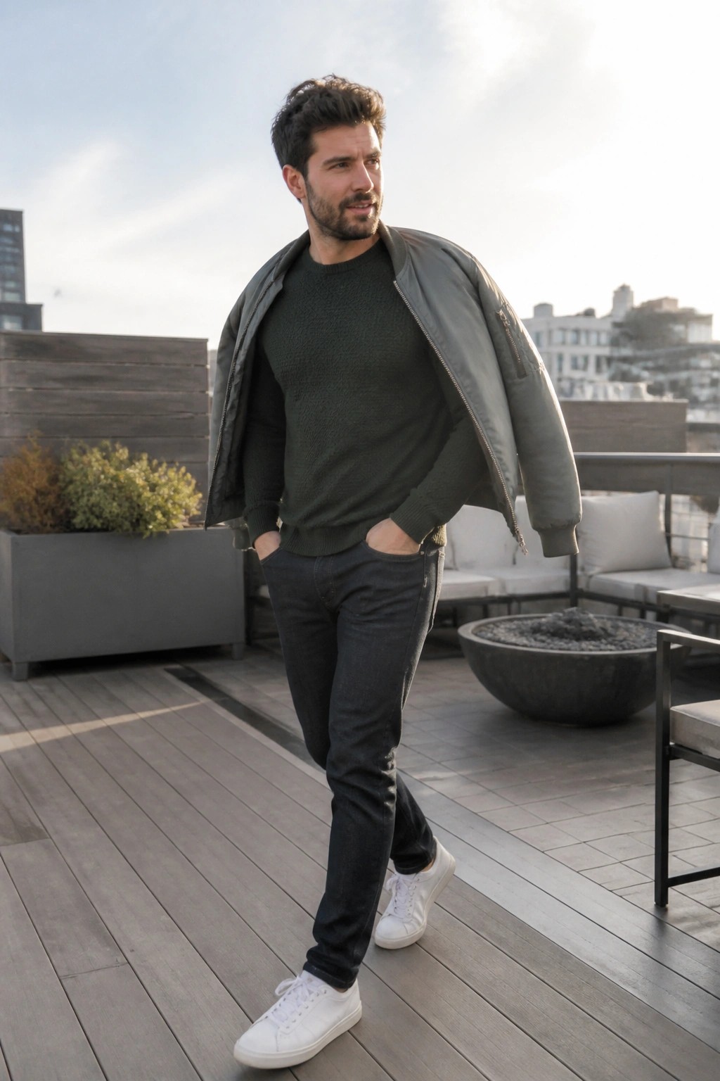 Man in green sweater and open grey jacket on sunny rooftop deck with city view.
