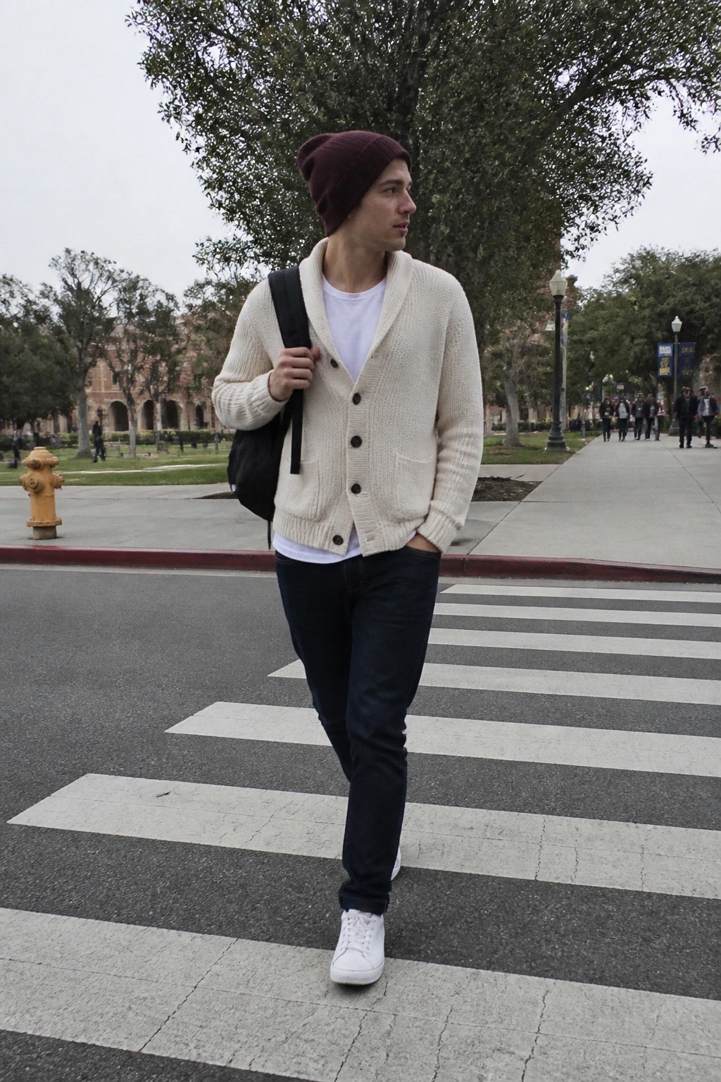 Young man in maroon beanie and cream cardigan crosses street with backpack