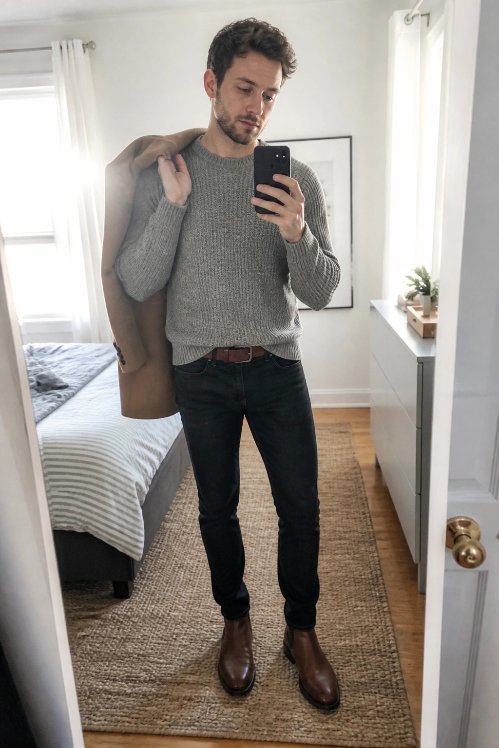 Man in gray sweater holds tan coat while taking bedroom mirror selfie with phone.