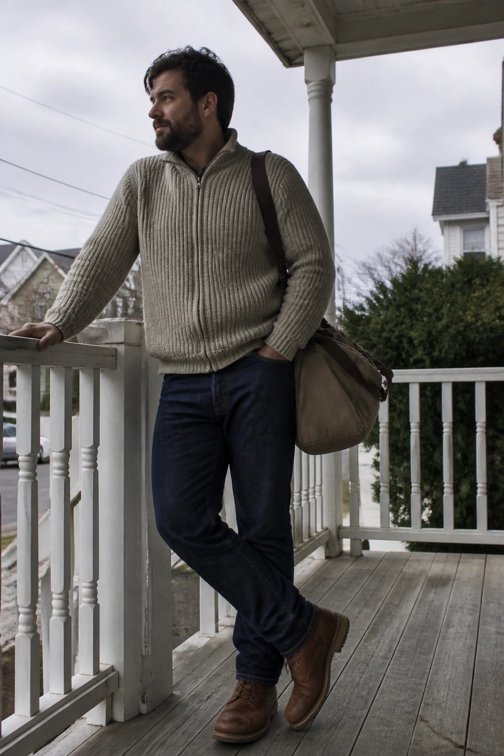 Man in beige zip sweater and jeans with bag on wooden porch leaning on railing