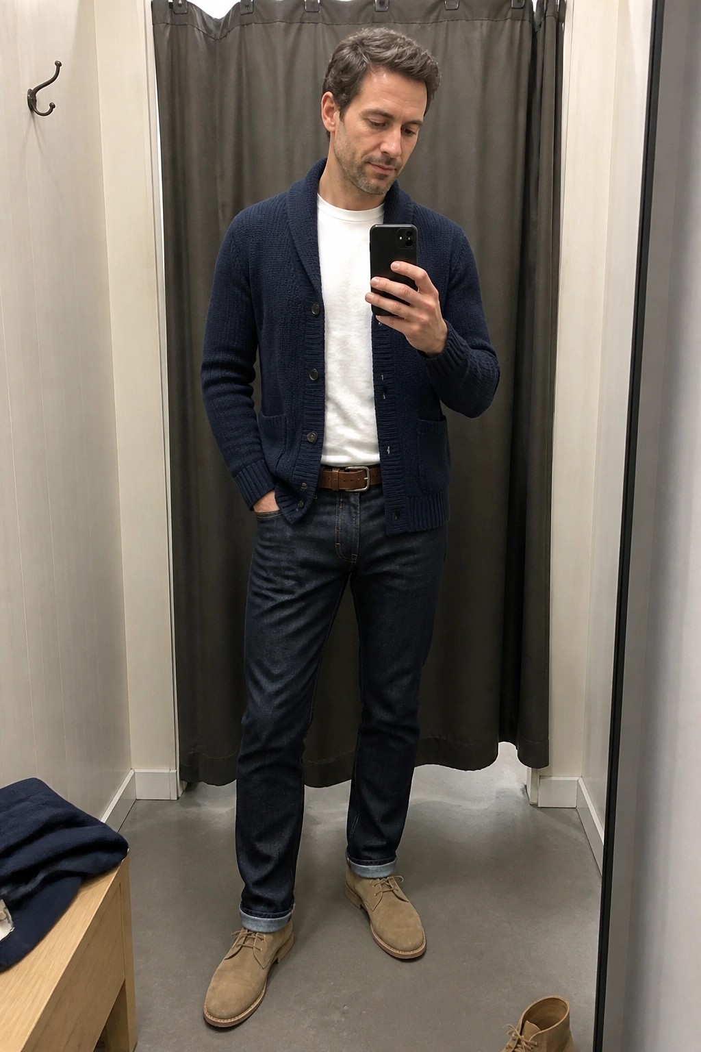 Man in navy cardigan and jeans takes mirror selfie in fitting room.