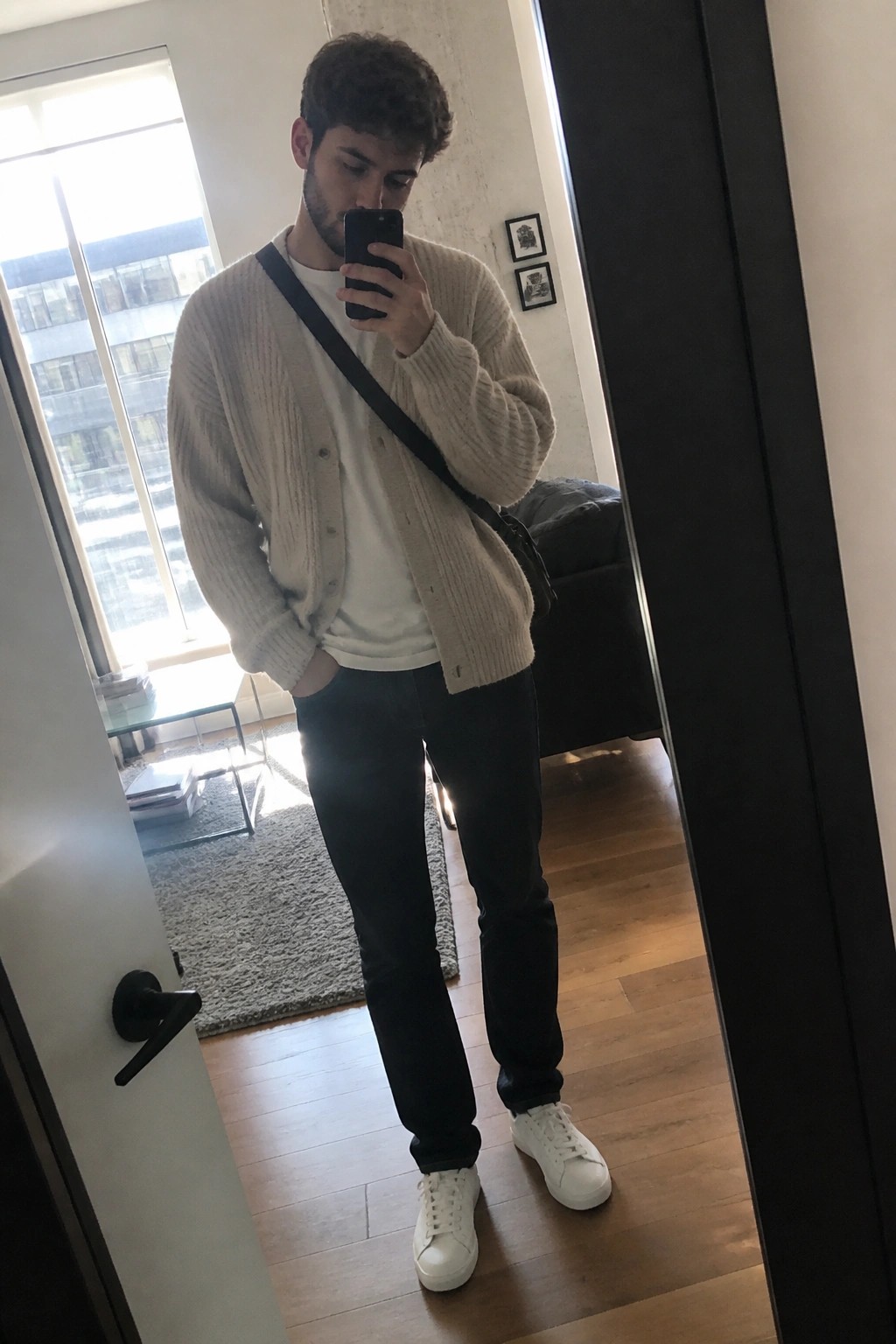 Man in beige cardigan over white tee takes mirror selfie in apartment