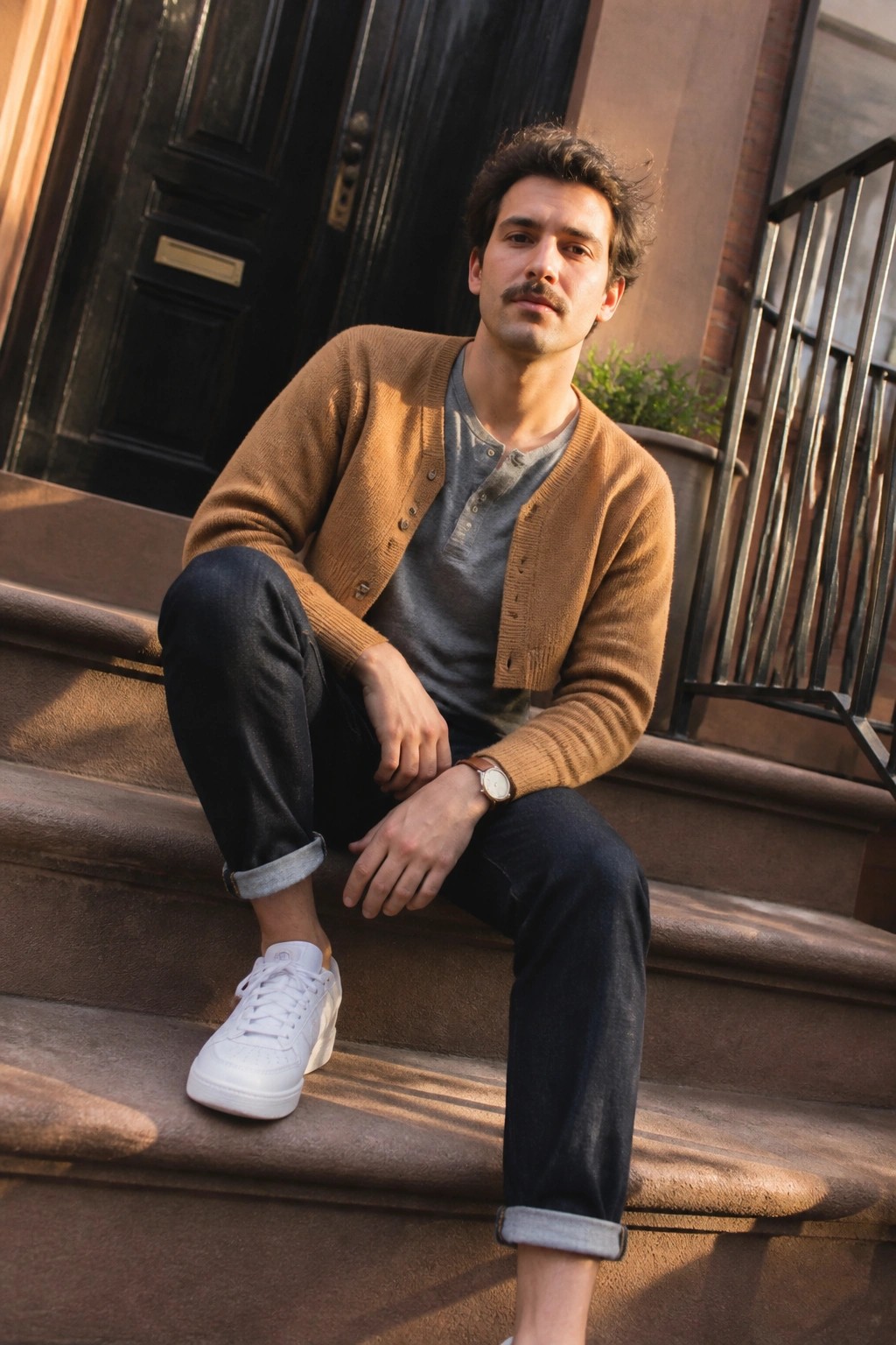 Man in brown cardigan, gray henley, dark jeans, and white sneakers on steps