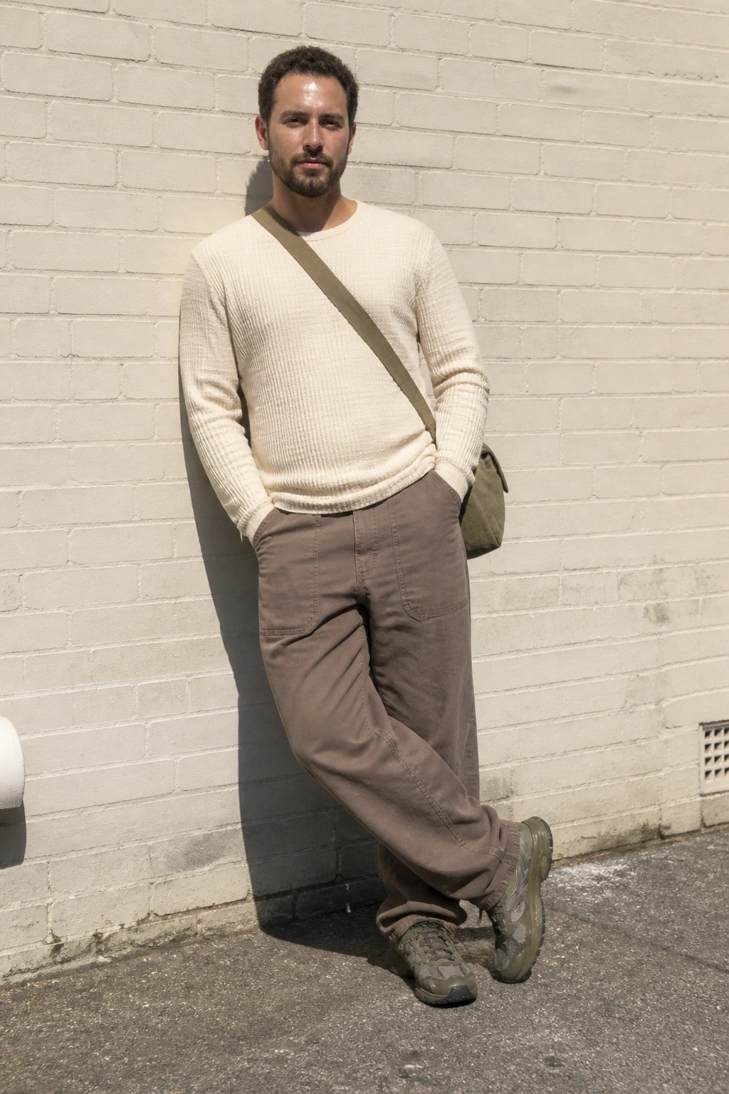 Man in cream sweater and brown pants with green bag leaning against white brick wall