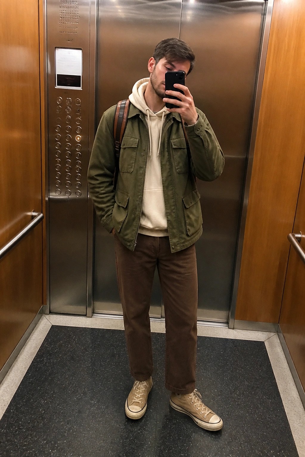 Man in green jacket over cream hoodie taking mirror selfie in elevator with brown pants