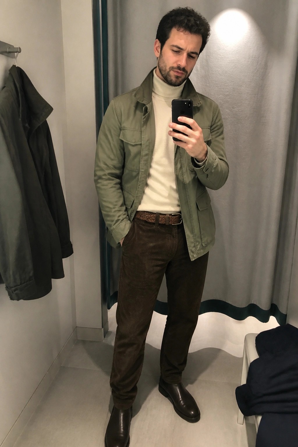 Man taking a mirror selfie in an olive green field jacket over a cream turtleneck sweater paired with brown corduroy pants a brown braided belt and dark brown leather boots.