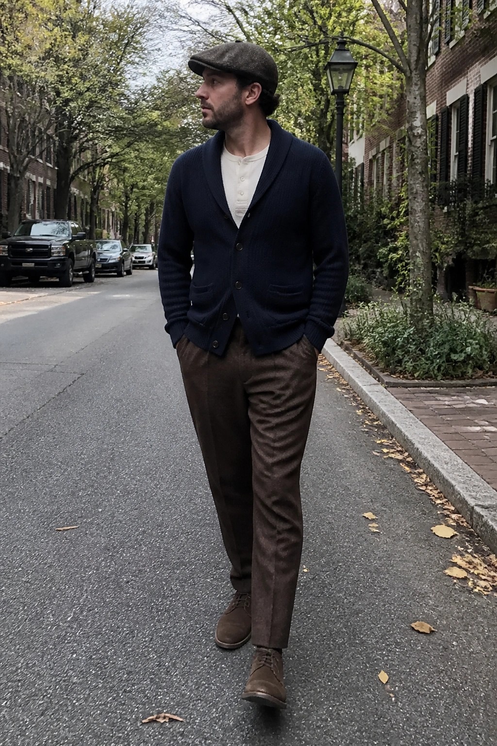 Man in brown flat cap, navy cardigan, and brown trousers on tree-lined street