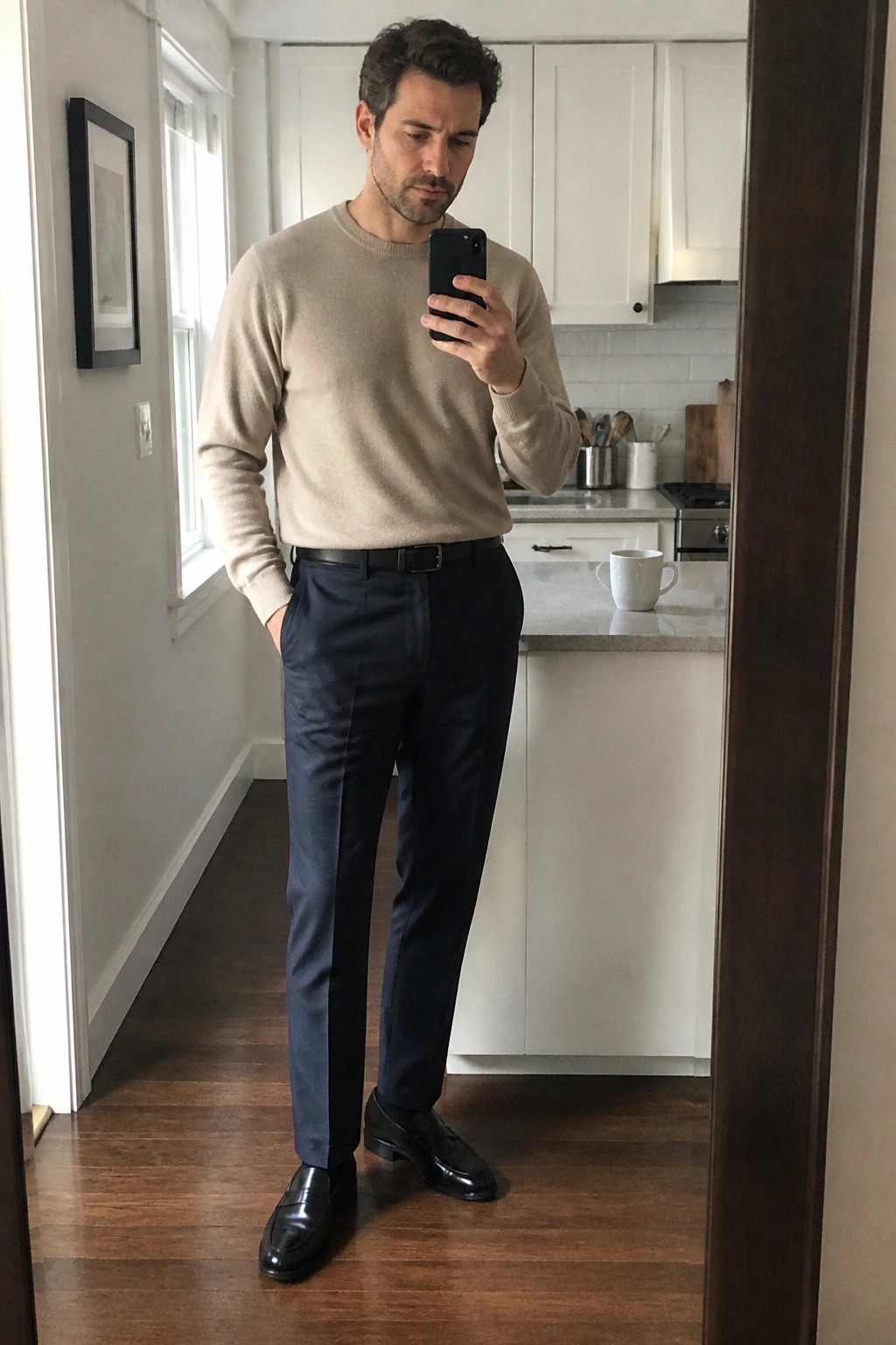 Man in beige crewneck sweater tucked into navy wool trousers with black belt and black leather loafers taking a mirror selfie indoors