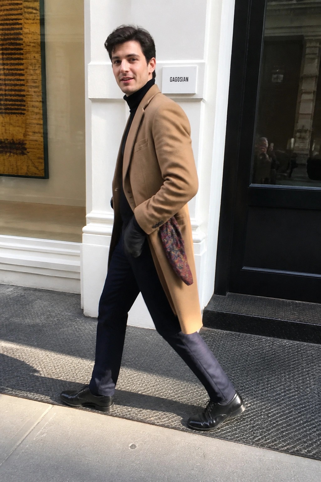 Man in a camel wool overcoat over a black turtleneck and navy wool trousers with black dress shoes and gloves, scarf tucked into the coat pocket.