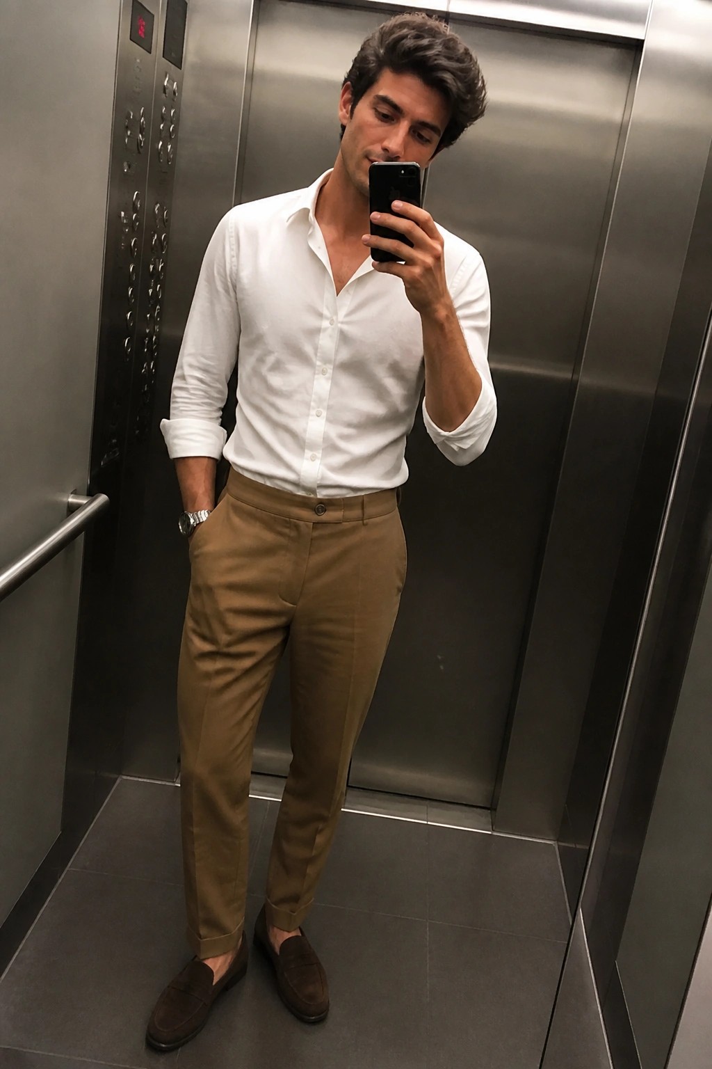 Man in white shirt and tan pants taking phone selfie in elevator