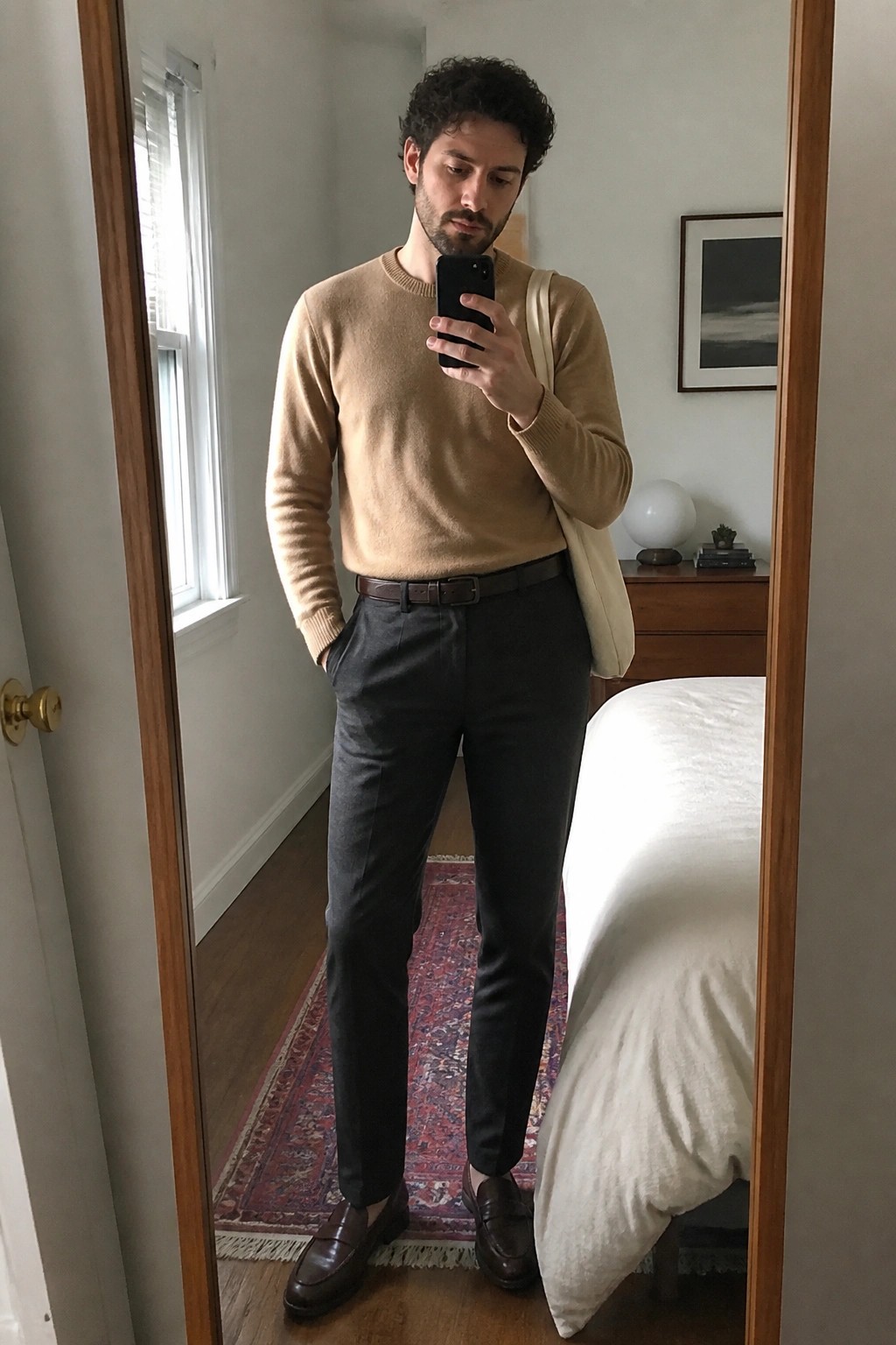 Curly-haired man in beige sweater and dark trousers taking bedroom mirror selfie.