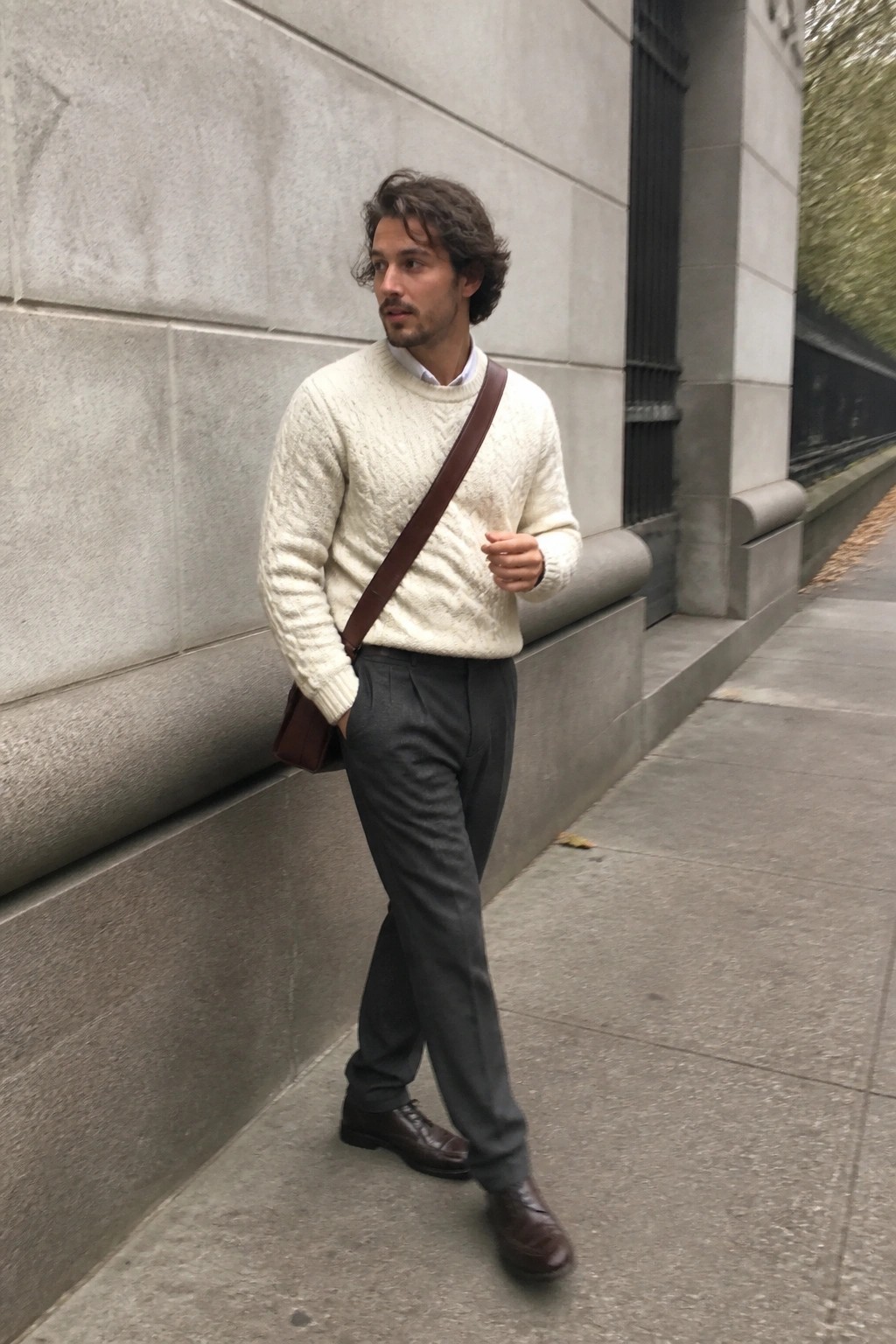 Man in cream cable-knit sweater, gray trousers, brown bag by stone wall