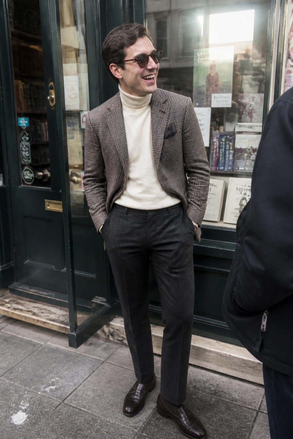 Man in checkered blazer and cream turtleneck smiles outside a bookstore.