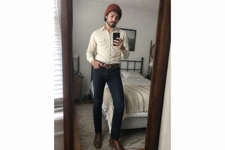 Man in cream embroidered shirt, rust beanie, jeans, and boots takes bedroom mirror selfie.