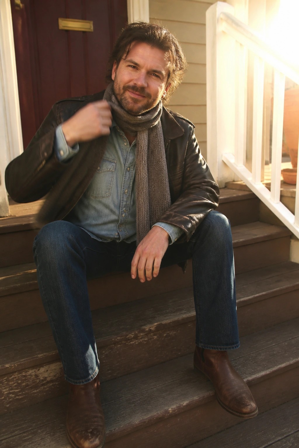 Bearded man in leather jacket adjusts scarf on sunlit wooden porch steps