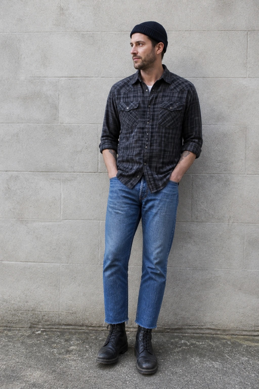 Man in black beanie, plaid shirt, and frayed jeans against concrete wall.
