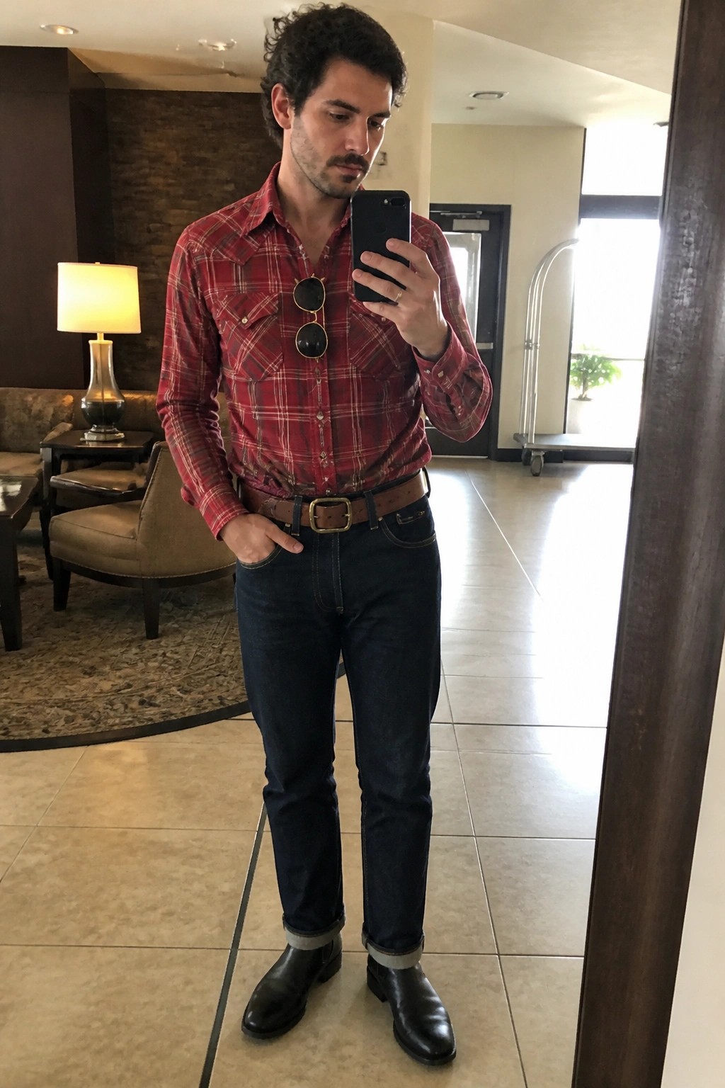 Man in red plaid shirt taking mirror selfie in hotel lobby wearing jeans and boots