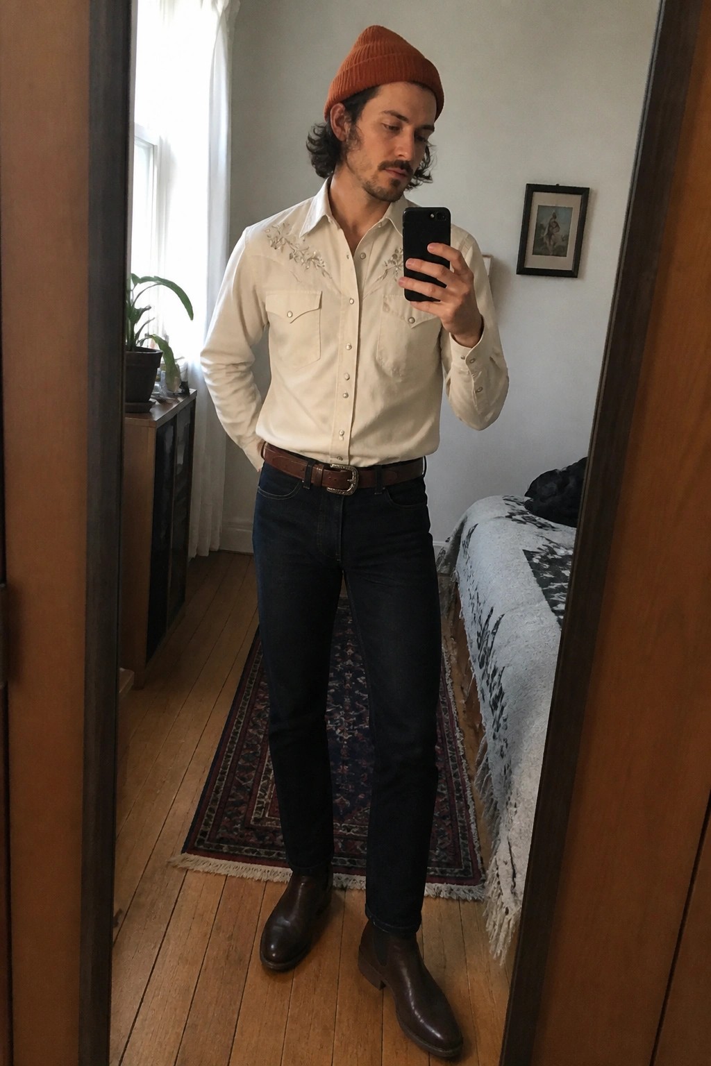 Man in cream embroidered shirt, rust beanie, jeans, and boots takes bedroom mirror selfie.