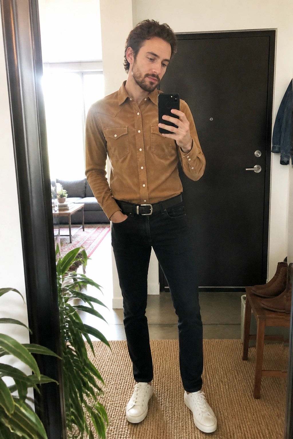 Bearded man in brown shirt, black jeans, and white sneakers taking mirror selfie