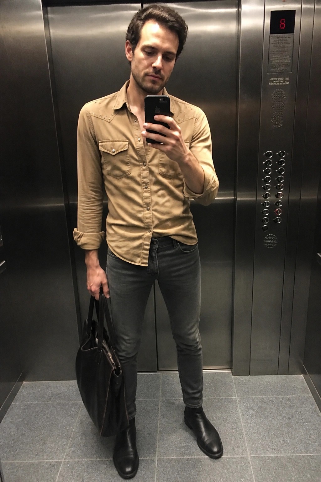 Man in tan studded shirt takes mirror selfie in elevator holding black tote bag