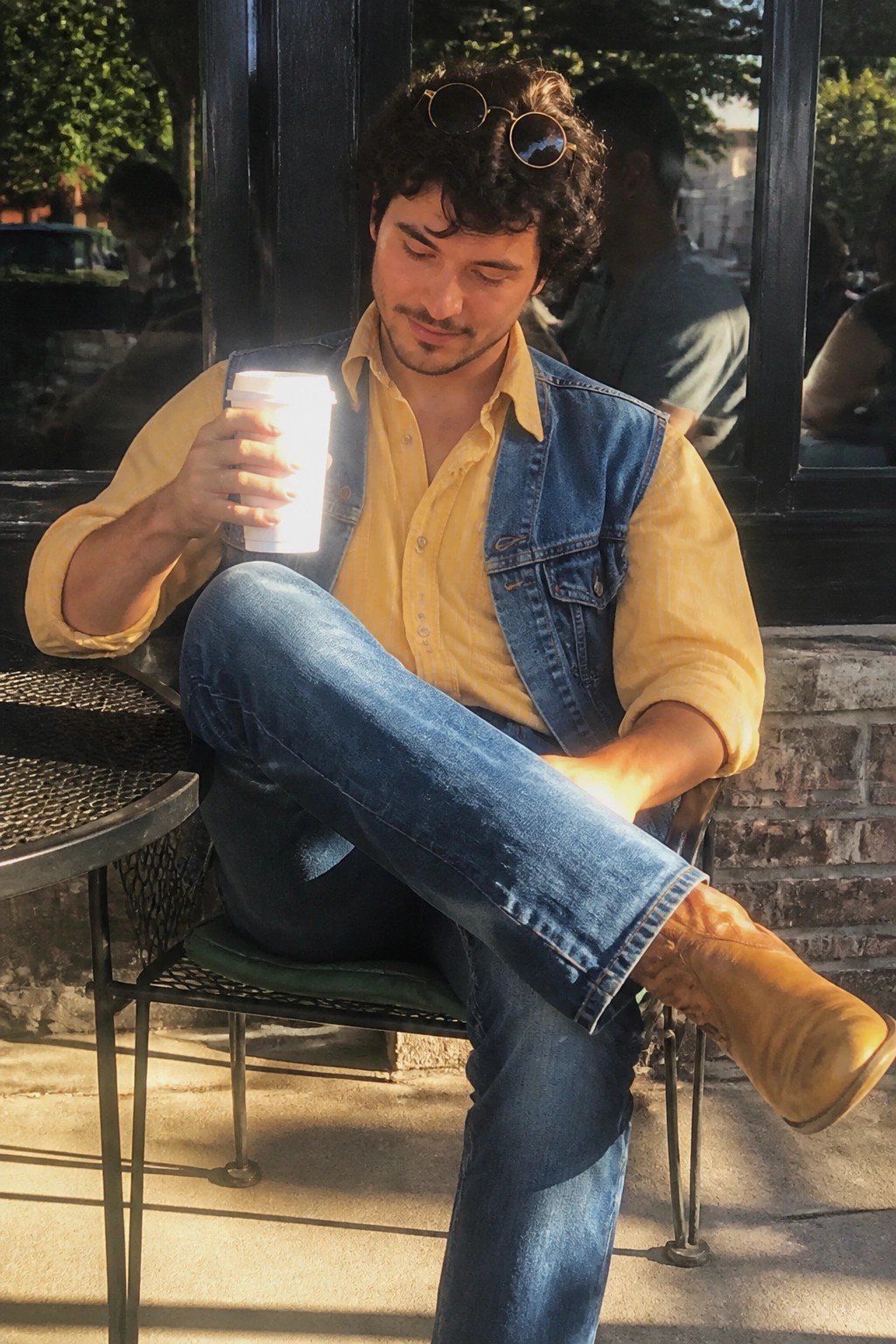 Young man in yellow shirt and denim vest holding coffee cup in sunlight