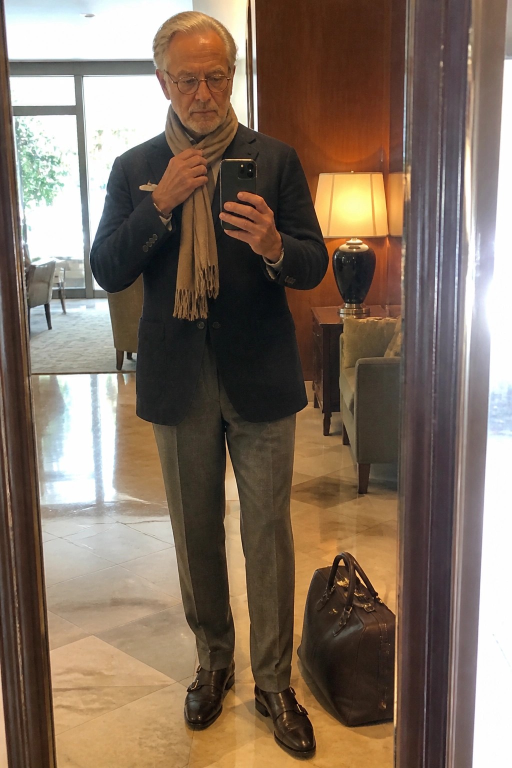 Elderly man in dark blazer and beige scarf takes mirror selfie beside leather bag