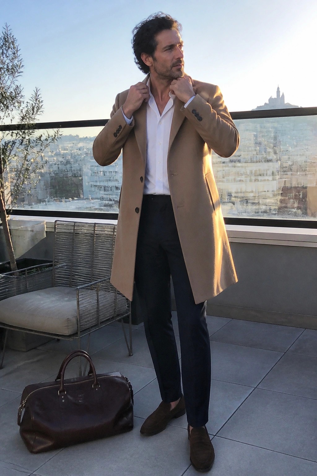 Man in tan overcoat over white shirt adjusts collar on city balcony with leather bag.