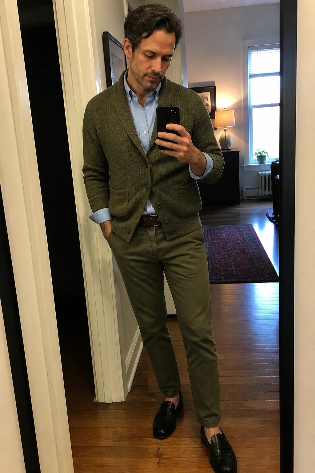 Man in green cardigan over blue shirt with olive pants holding smartphone in mirror
