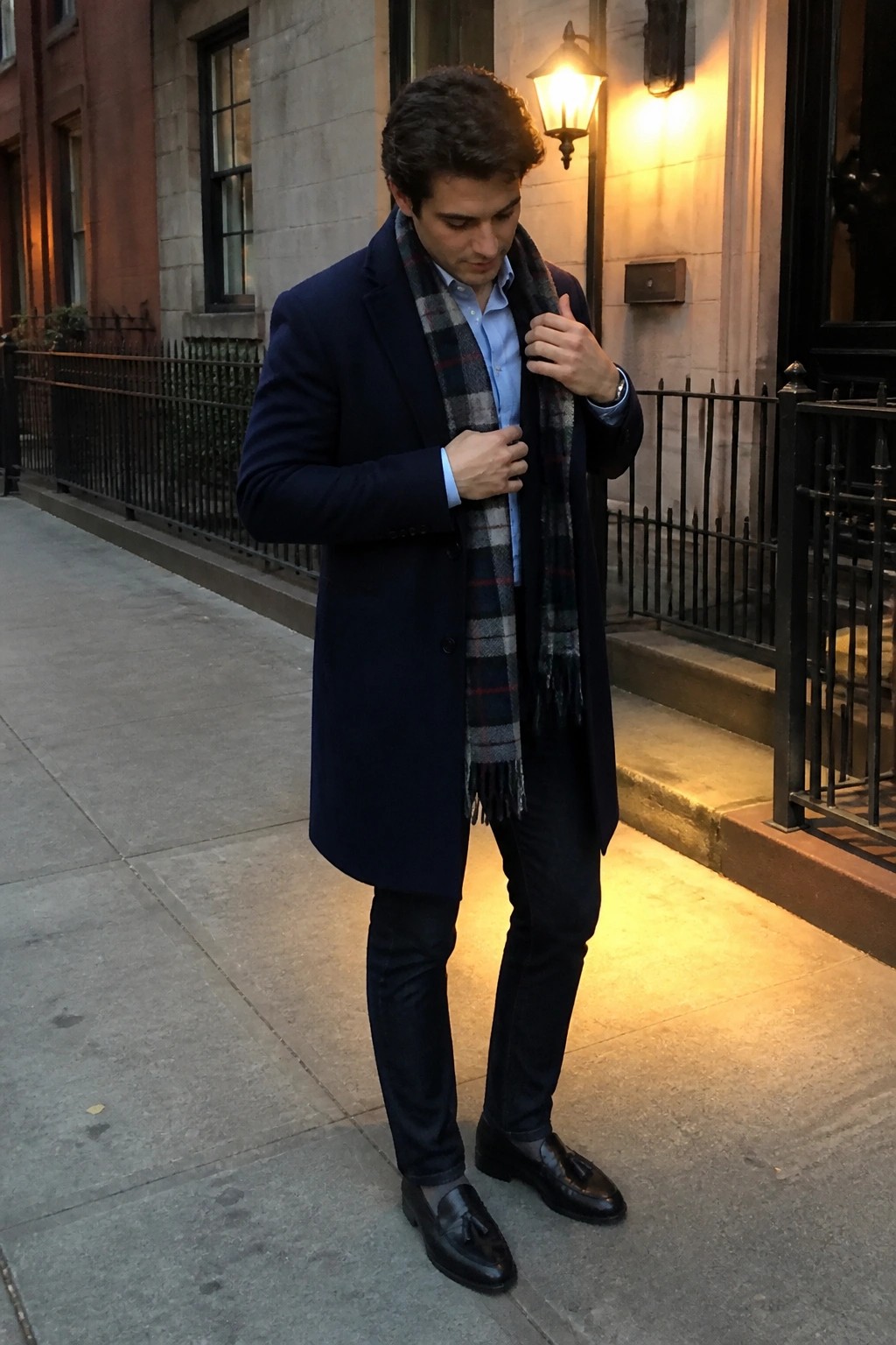 Man in navy overcoat adjusts plaid scarf on city sidewalk at dusk