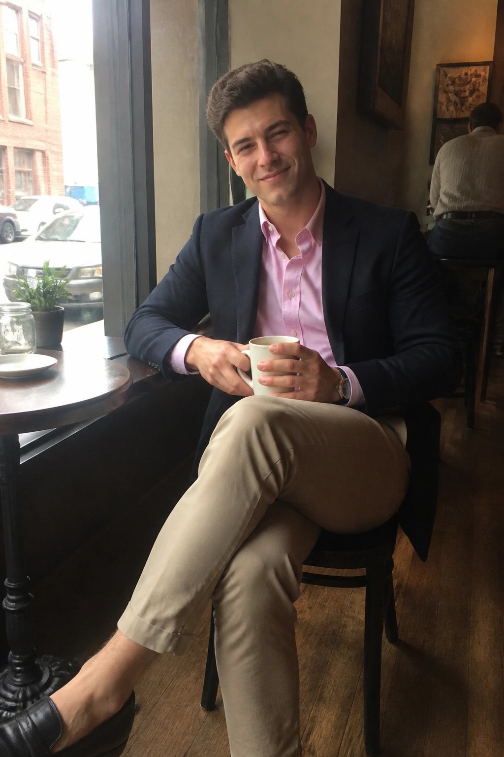 Man seated in cafe wearing navy blazer over pink oxford shirt, khaki pants, and black loafers while holding a white mug.