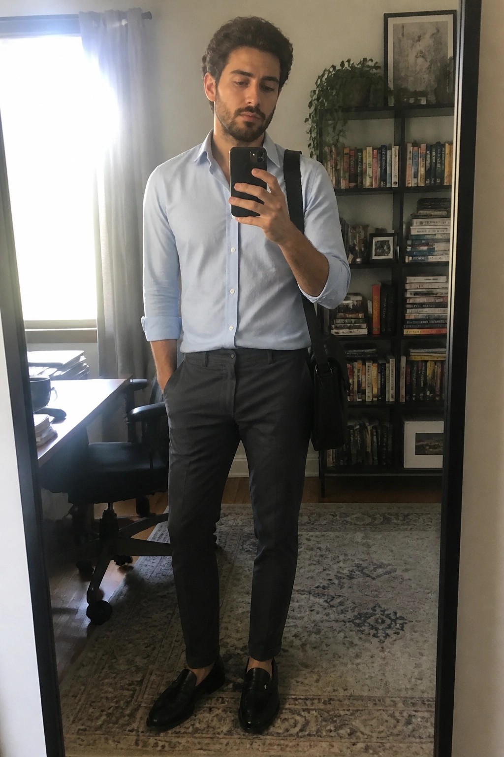 Man in light blue shirt and gray pants taking mirror selfie before bookshelves